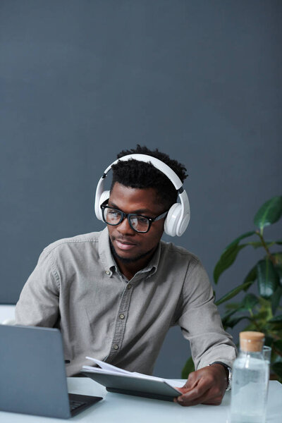 Vertical image of African American man in headphones sitting at table with laptop and working online