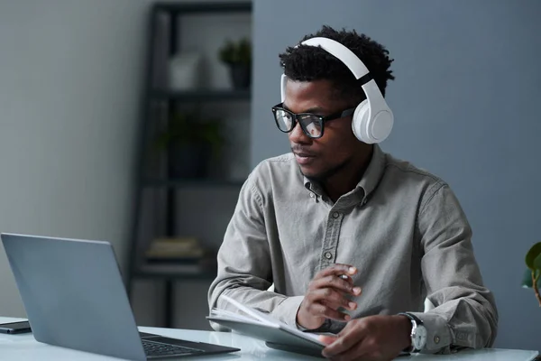 African American businessman in wireless headphones looking at monitor of laptop sitting at his workplace, he doing his online work