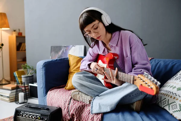 坐在房间沙发上的年轻女子戴着无线耳机 专心致志地弹吉他 — 图库照片