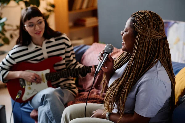 非洲裔美国女孩在麦克风里唱歌 而她的朋友在弹吉他 她们坐在房间的沙发上 — 图库照片