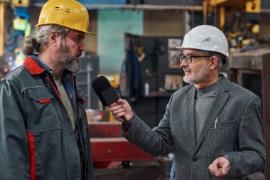 Üniformalı, olgun sakallı bir işçi fabrikada röportaj sırasında TV muhabiriyle konuşuyor.