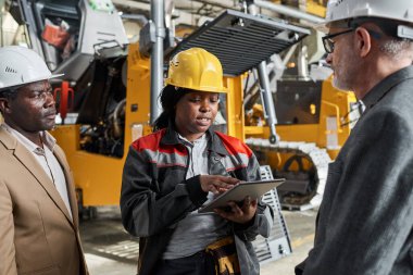 Üniformalı Afrikalı kadın işçiler tablet bilgisayar kullanıyor ve raporunu mühendislere sunuyor. Depoda arka planda makinelerle duruyorlar.