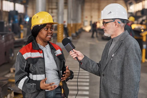 News reporter in work helmet holding microphone in front of the worker and interviewing her during her work in factory