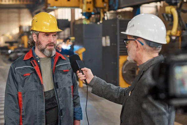 Mature worker in uniform giving an interview to microphone for reporter while they standing in workshop in factory