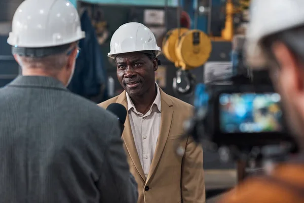 African confident engineer in hardhat talking to reporter during online broadcasting in factory