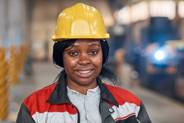Portrait of African young woman in work helmet and uniform smiling at camera working in factory
