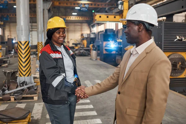 African manager in suit shaking hands with female worker in uniform to hire her in factory