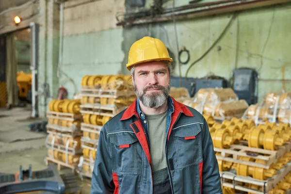 Portrait of mature manual worker in work helmet and uniform looking at camera while working in warehouse