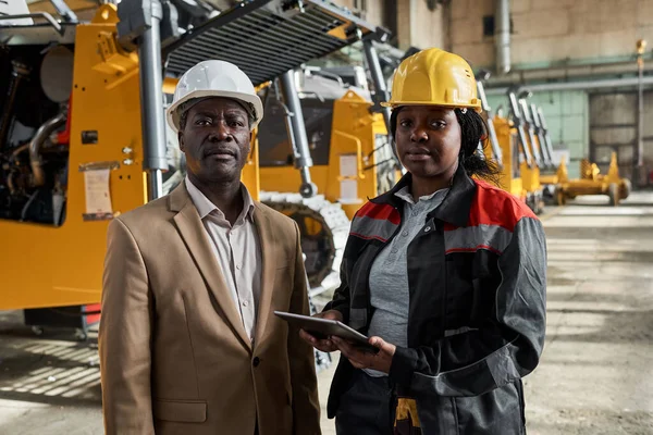 Portrait of African engineer with female mechanic standing in factory and looking at camera while working online on tablet pc