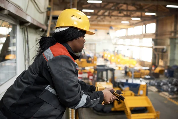African female worker in uniform and hardhat standing on balcony and watching at empty warehouse with machines