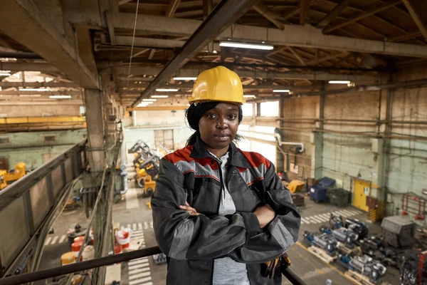 Portrait of African female worker in work helmet and uniform standing on balcony with her arms crossed and looking at camera