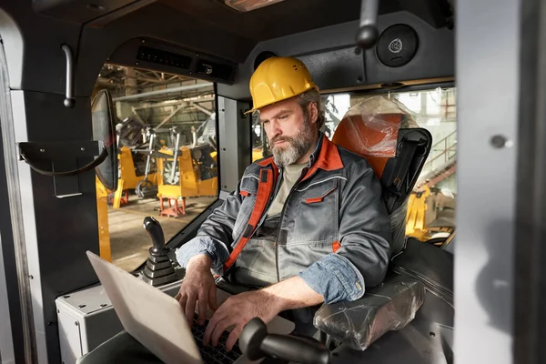 Mature operator in uniform and work helmet sitting in cabin of construction machine and controlling the work with laptop