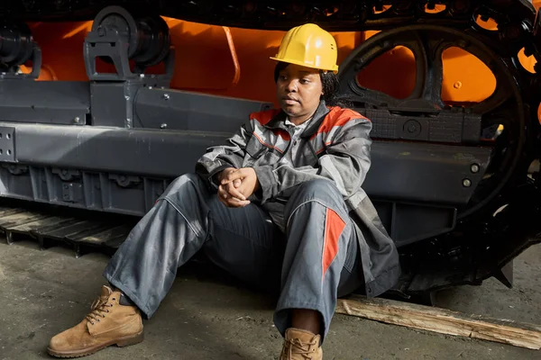 African female worker in uniform and hardhat sitting on the floor leaning on tractor and resting after work