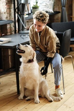 Protez kollu genç bir kadın av köpeğini okşuyor ve sandalyede otururken gülümsüyor.