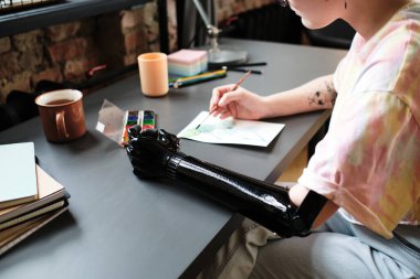 Protez kollu genç bir kadının yakın plan fotoğrafı. Elinde kağıtla masada oturuyor ve boya fırçasıyla resim yapmayı öğreniyor.