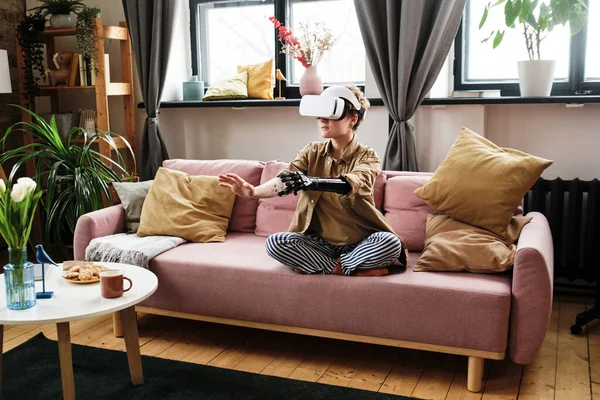 Girl with prosthetic arm in goggles sitting on sofa and gesturing while playing virtual reality game in living room