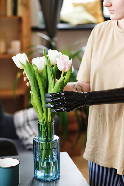 Close-up of young woman with prosthetic arm putting white tulips in vase with water on table