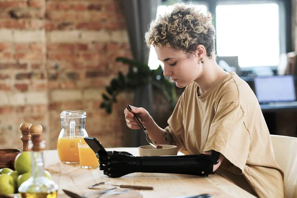 Young woman with prosthetic arm using her mobile phone while having breakfast at table