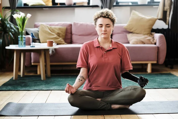 Young woman with prosthetic arm sitting on exercise mat in living room in lotus position and meditating with her eyes closed