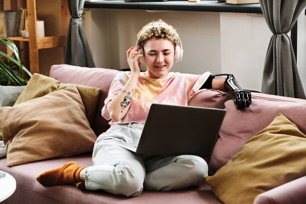 Happy young woman with prosthetic arm wearing wireless headphones talking online using her laptop on sofa in room