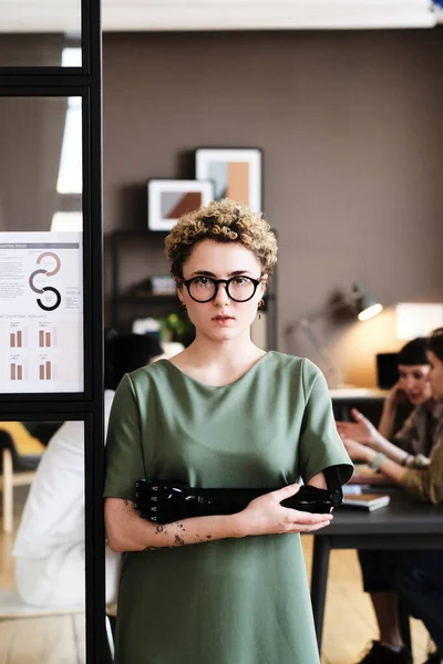 Portrait of young woman in eyeglasses with prosthetic arm looking at camera while standing at office with colleagues behind her