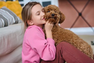 Küçük kız sevimli köpeğine sarılıp evdeki odada onunla oynuyor.