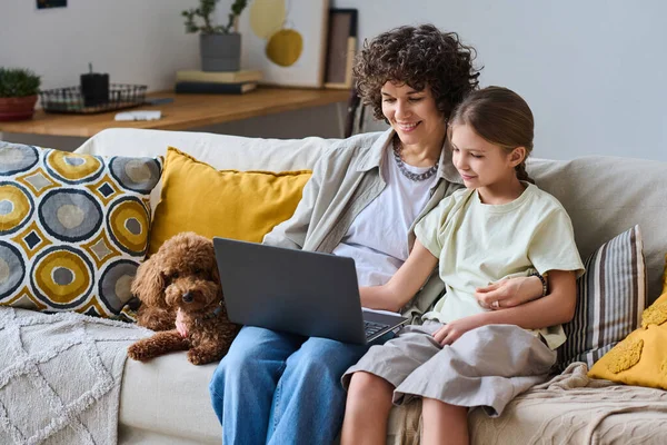 Mom and daughter using laptop together while sitting on sofa with their dog at home