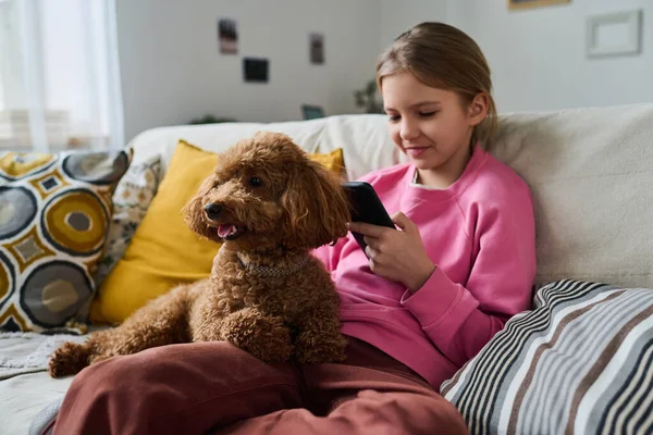 Little girl sitting on sofa with dog and playing game in her smartphone during leisure time at home