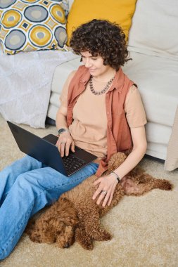 Evde köpekle dinlenen ve dizüstü bilgisayar kullanan genç bir kadının dikey görüntüsü