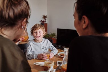 Küçük mutlu çocuk meyve suyu içiyor ve yemek saatinde masada lezbiyen anneleriyle konuşuyor.