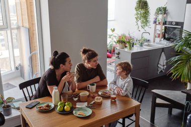 Lezbiyen anneler mutfakta kahvaltı yaparken yemek yiyip oğluyla konuşurlar.