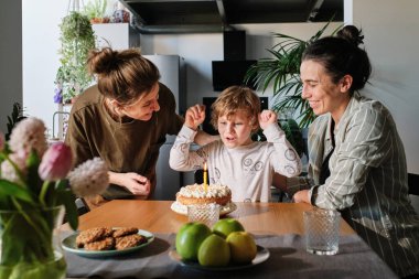 Küçük çocuk elinde mumla oturup homoseksüel anneleriyle doğum gününü kutlarken dilek tutuyor.