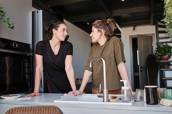 stock image Young lesbian couple looking at each other while standing at table with sink during household in kitchen