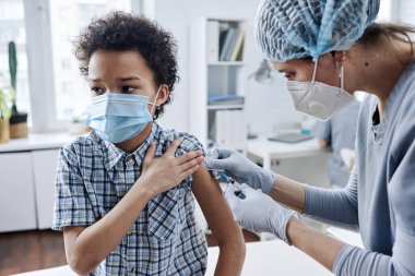 Kolunu aşılamak için şırınga kullanan küçük Afrikalı bir çocuk doktor muayenehanesinde oturuyor.