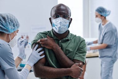 Maskeli Afrikalı bir hastanın portresi. Doktor omzuna aşı yaparken kameraya bakıyor.