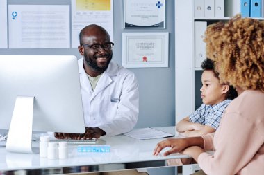 Ofisteki bilgisayarda reçete yazarken Afrikalı erkek doktor küçük bir hastaya ve annesine gülümsüyor.