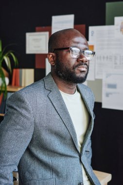 Gözlüklü Afrikalı ciddi işadamı ofiste dikilirken düşünceli görünüyor.