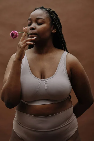 Portrait of African plump woman in underwear holding rose flower in her mouth isolated on brown background
