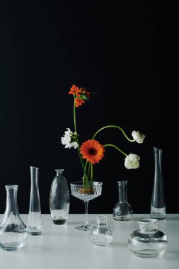 Dikey çağdaş renkli çiçeklerin ve gri masa yüzeyindeki çeşitli şeffaf cam damarların siyah duvar arka planına karşı yaşam bileşimi