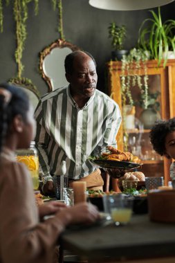 Afrikalı olgun bir adam yemek masasında ailesiyle yemek yerken servis ediyor.