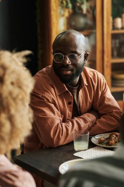 Gözlüklü Afrikalı genç adam yemek sırasında masada bir kadınla konuşuyor.
