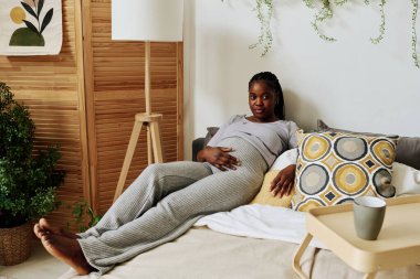 Yatağında uzanıp dinlenirken hamile karnına dokunan Afrikalı bir kızın portresi.