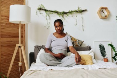 Yatak odasında boş zamanlarında yatakta oturmuş kameraya bakan Afrikalı hamile bir kadının portresi.