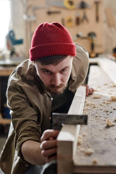 Vertical shot of serious young male carpenter using steel square when working with wood plank in woodshop