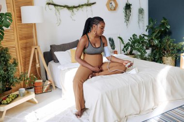 Afrikalı hamile bir kadın sağlığını önemsiyor. Yatak odasında oturup kan basıncını ölçüyor.