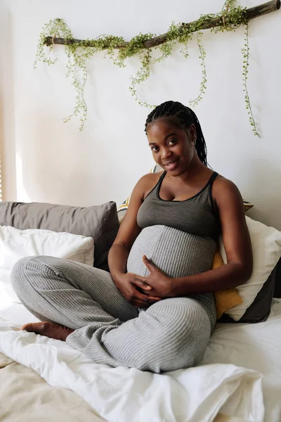 非洲怀孕女孩在床上休息时触摸怀孕的腹部和对着相机微笑的画像 — 图库照片