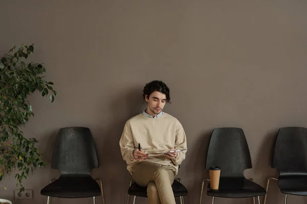 Młody Człowiek Siedzi Krześle Korytarzu Czyta Swoje Czekając Wywiad — Zdjęcie stockowe
