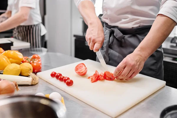 テーブルでまな板に新鮮なトマトをカットするナイフを使用してエプロンで若い料理人のクローズアップ — ストック写真