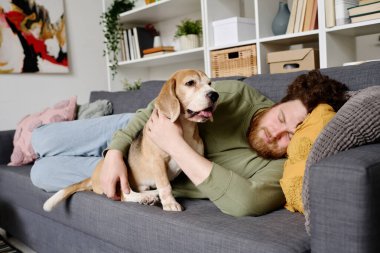 Sahibi oturma odasında kanepede uzanıyor ve yanında oturan sevimli köpeğiyle yatıyor.