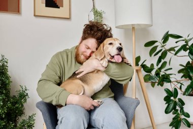 Genç sakallı adam, salonda koltukta oturmuş köpeğini kucaklayan ve rahatlatan bitkilerle.